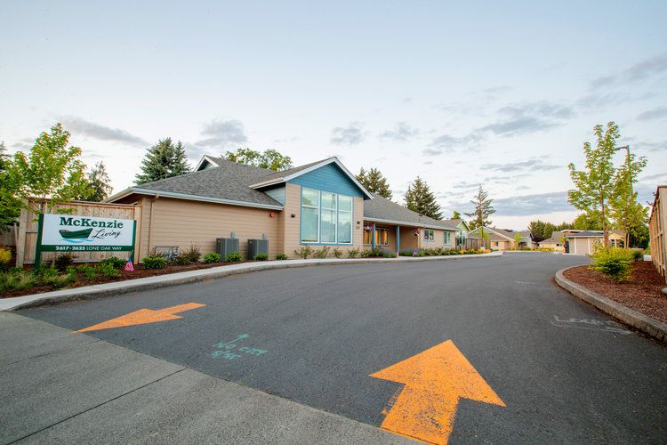 A modern building with a parking lot featuring directional arrows, surrounded by trees under a cloudy sky.