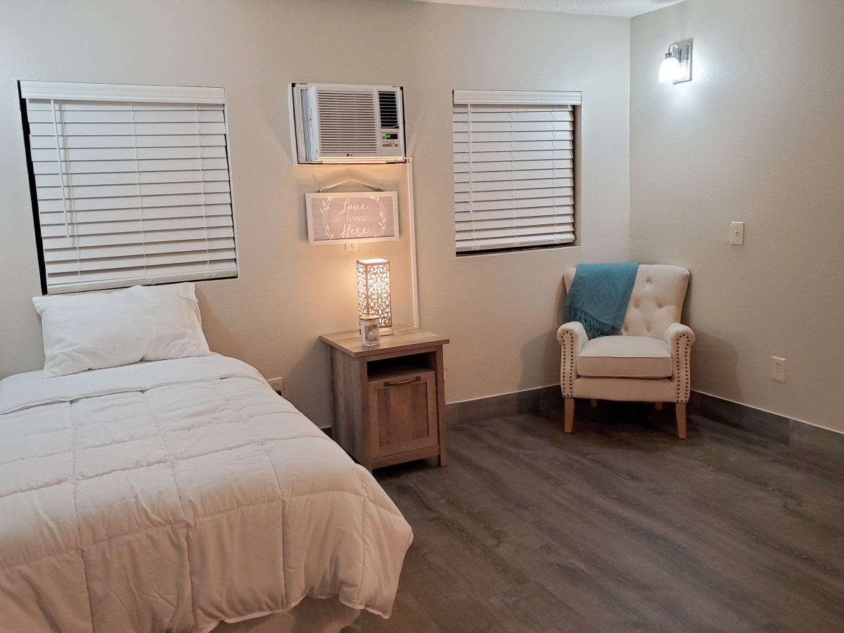 A cozy bedroom with a bed, nightstand, and armchair, featuring warm lighting and an air conditioner.