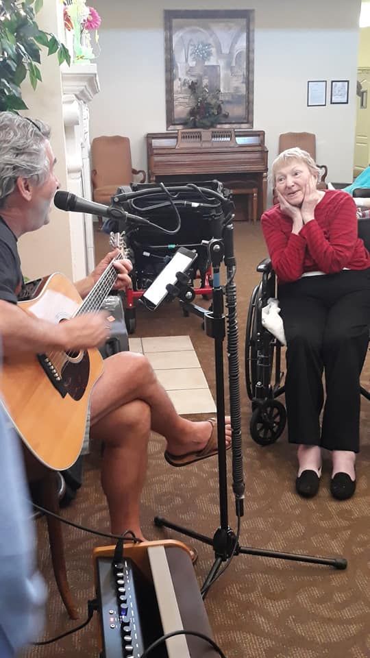 A musician plays the guitar and sings to an elderly woman in a wheelchair, creating a joyful moment.