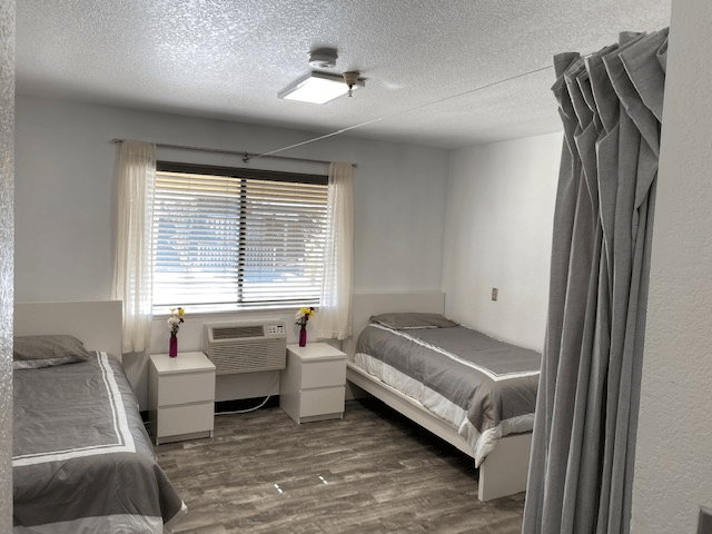 A minimalist bedroom with two single beds, nightstands, and an air conditioner beneath a window.