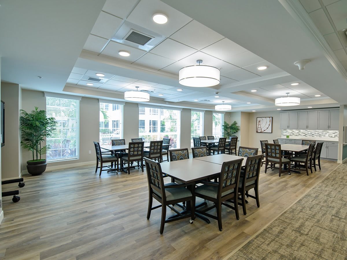 A spacious and well-lit dining area with multiple tables and chairs, featuring large windows and wooden flooring.