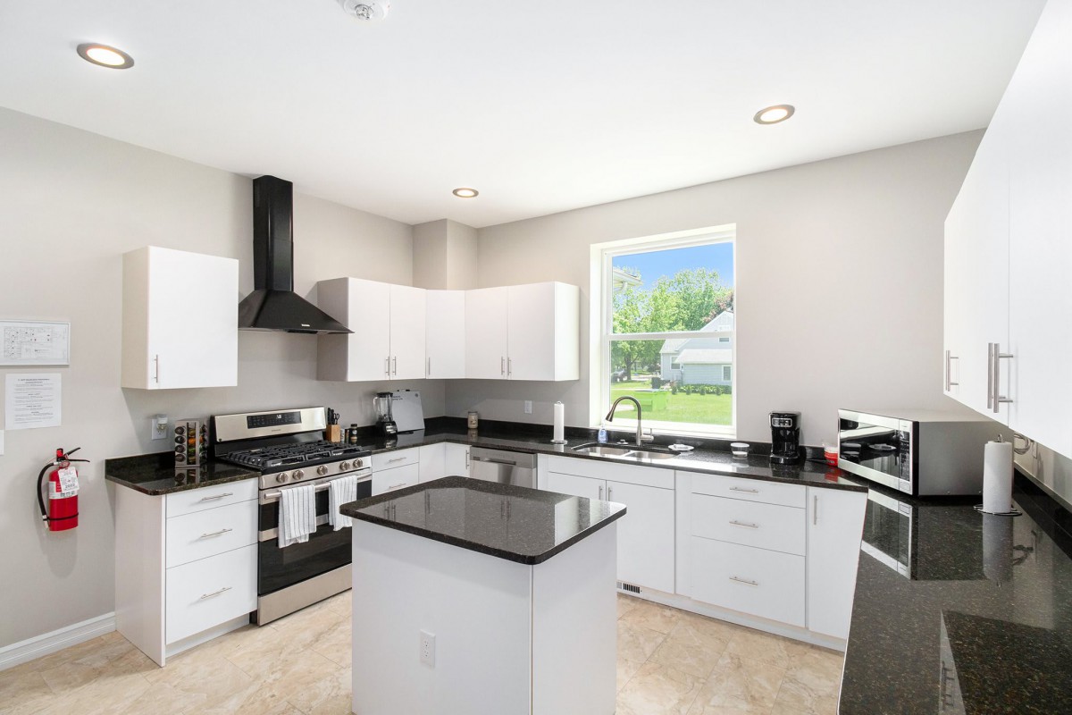 A modern kitchen with white cabinets, a central island, and stainless steel appliances, illuminated by natural light from a large window.