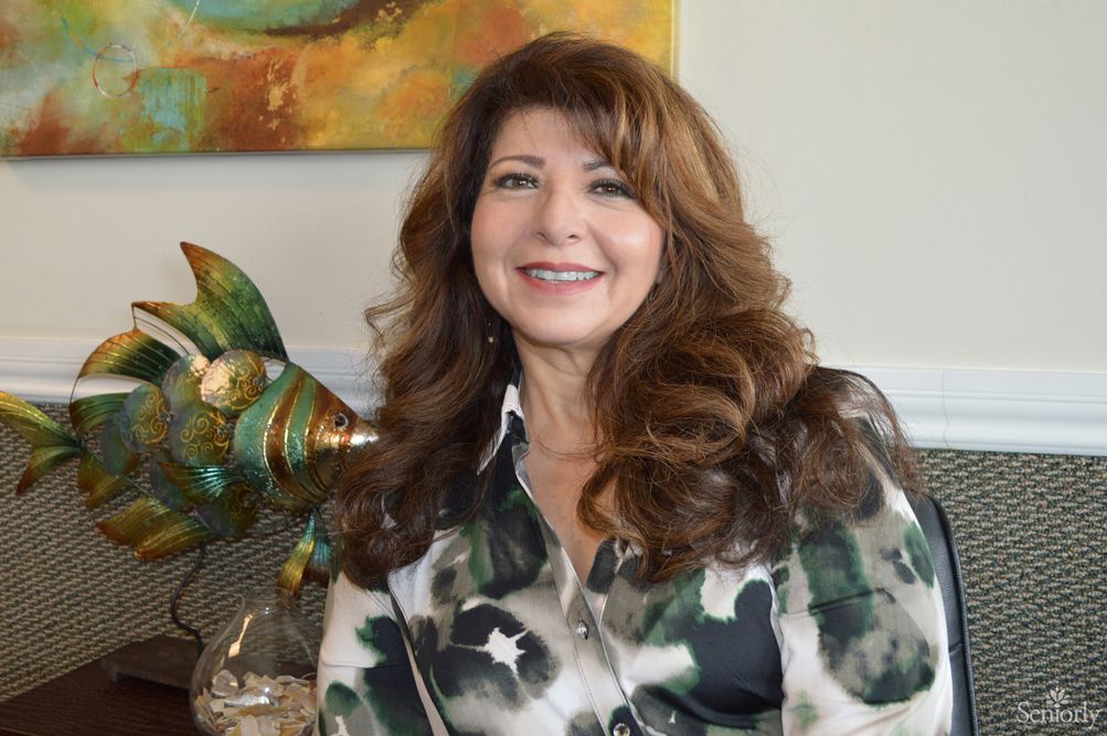 A woman with long hair is smiling indoors, seated next to decorative items and a painting in the background.