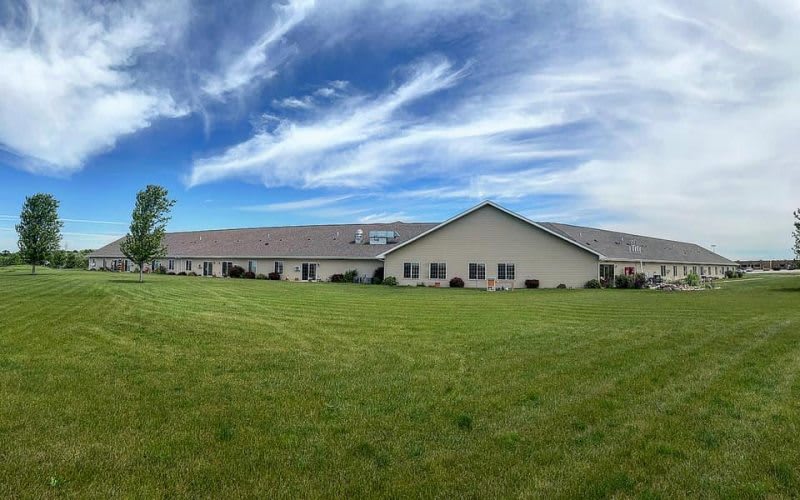 A wide-angle view of a single-story building surrounded by a grassy field under a blue sky with clouds.