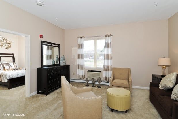 A cozy living room with two chairs, a round ottoman, and a large mirror near the window.