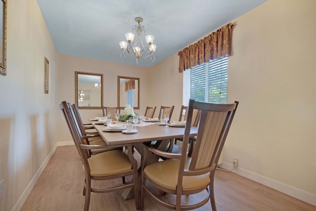 A well-lit dining room with a large table and eight chairs, illuminated by a chandelier, featuring natural light from a window.