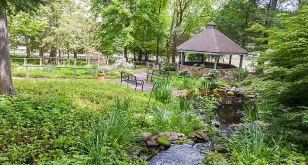 A serene garden featuring a gazebo, benches, a flowing stream, and lush greenery.