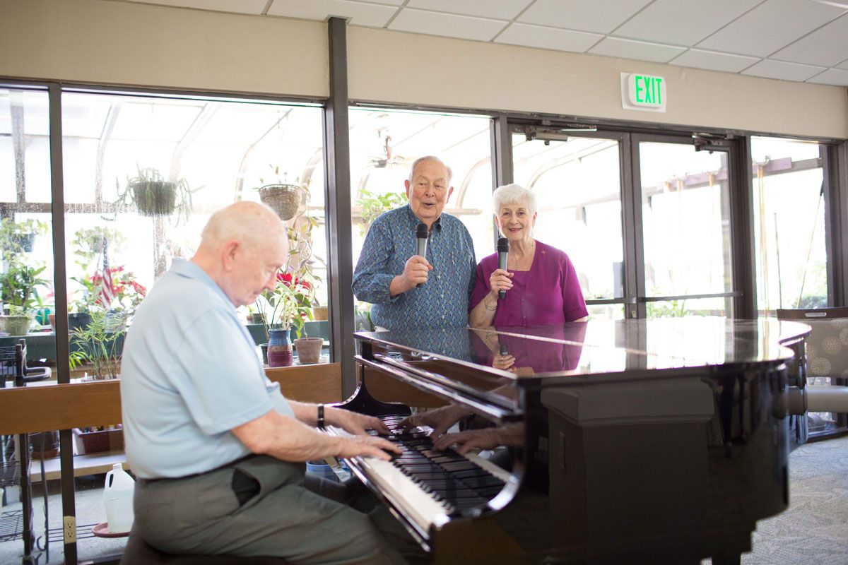 Elderly individuals enjoying music together, with one playing the piano and two others singing.