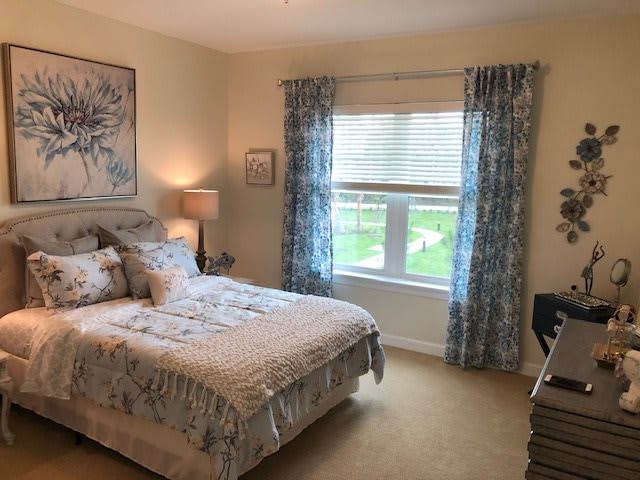 A cozy bedroom with a neatly made bed, curtains framing a large window, and decorative elements like lamps and artwork.