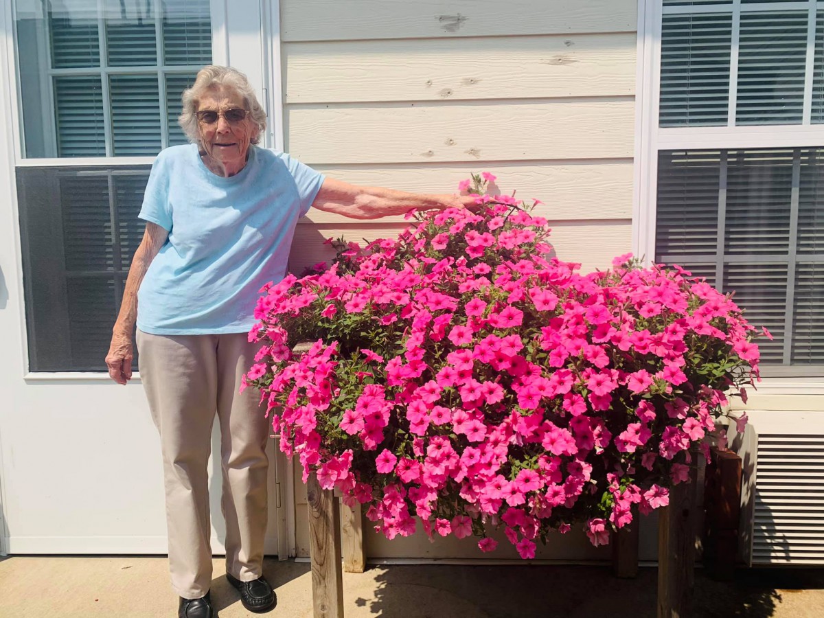 A woman stands beside a vibrant display of pink flowers on a sunny day.