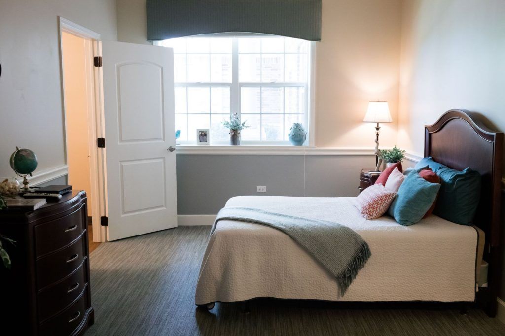 A modern bedroom interior with a neatly made bed, ambient lighting, and natural light streaming through a large window.