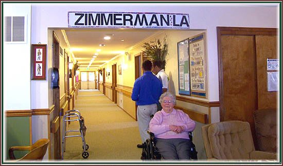 An elderly woman in a wheelchair sits in a hallway of a nursing home, with signage reading 'Zimmerman LA'.