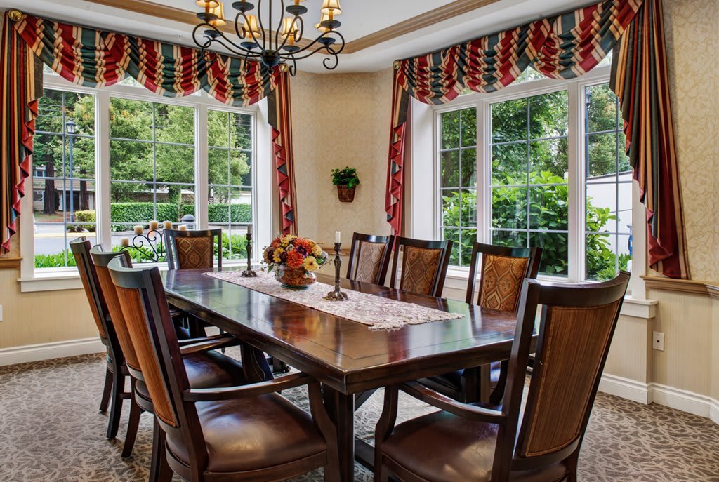 A dining room with a wooden table, surrounded by chairs and adorned with a chandelier, featuring large windows with decorative curtains.