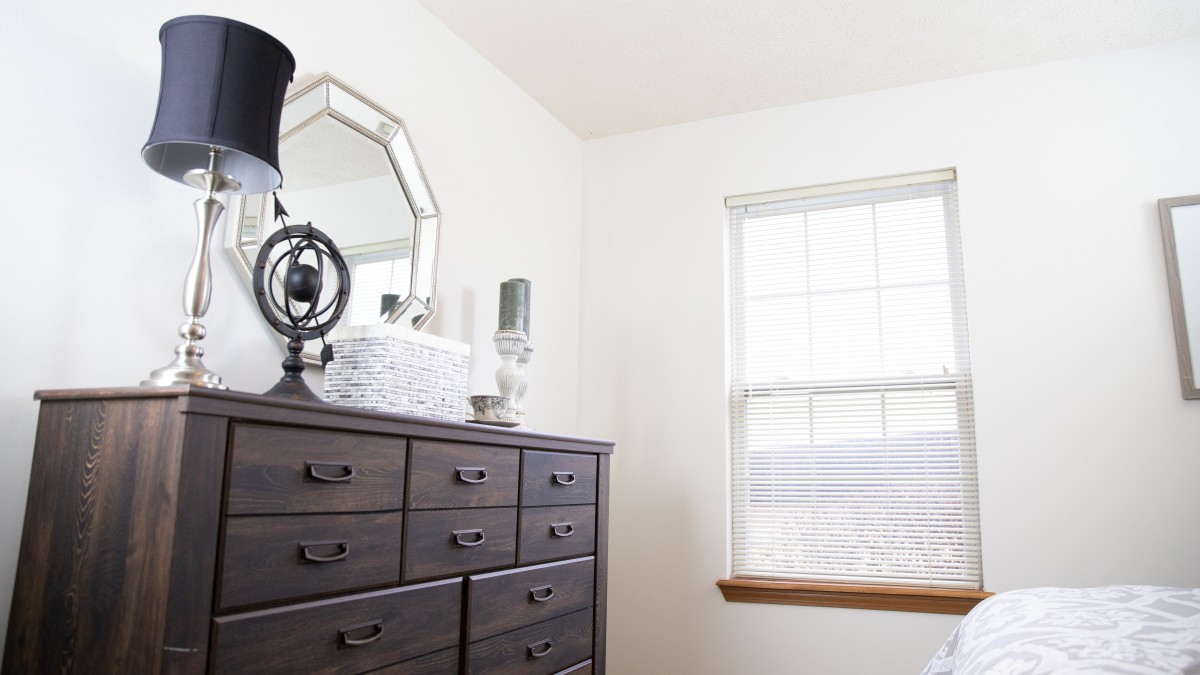 A bedroom dresser with decor items, including a mirror and a lamp, beside a window.