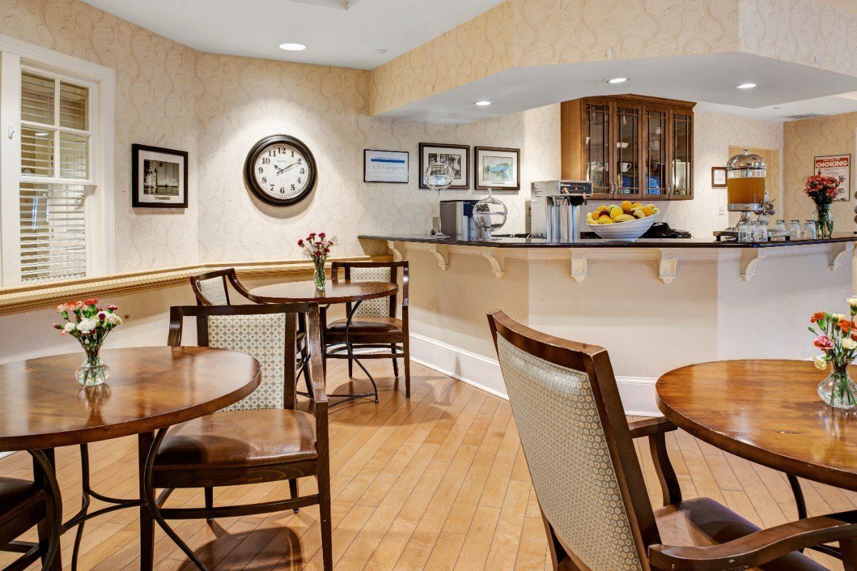 A cozy cafe interior featuring several tables and chairs, a large wall clock, framed pictures, and a coffee station with a wooden floor.
