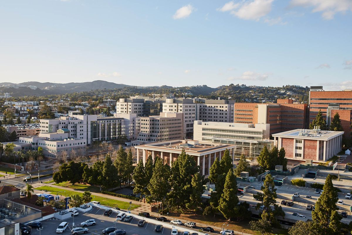 A scenic view of a university campus with multiple buildings, surrounded by greenery and hills under a clear sky.