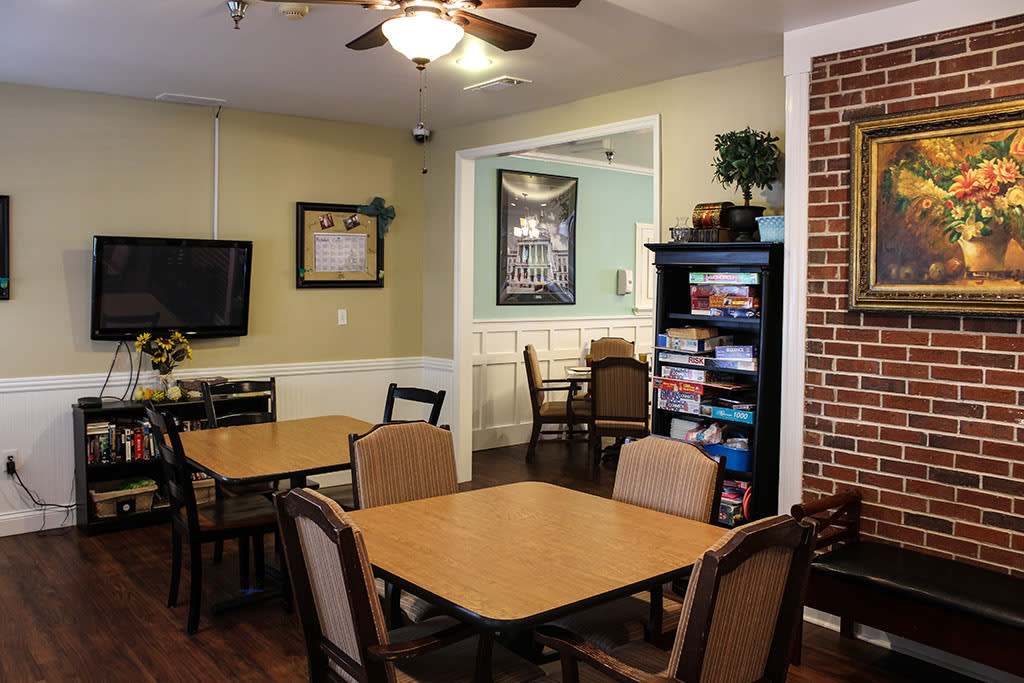 A cozy dining area with wooden tables and chairs, featuring a bookshelf and a brick accent wall.