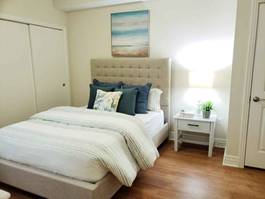 A cozy bedroom with a neutral color scheme, featuring a tufted headboard, decorative pillows, and a nightstand with a lamp.