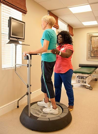 A physical therapist assists a patient on a balance training machine in a rehabilitation setting.