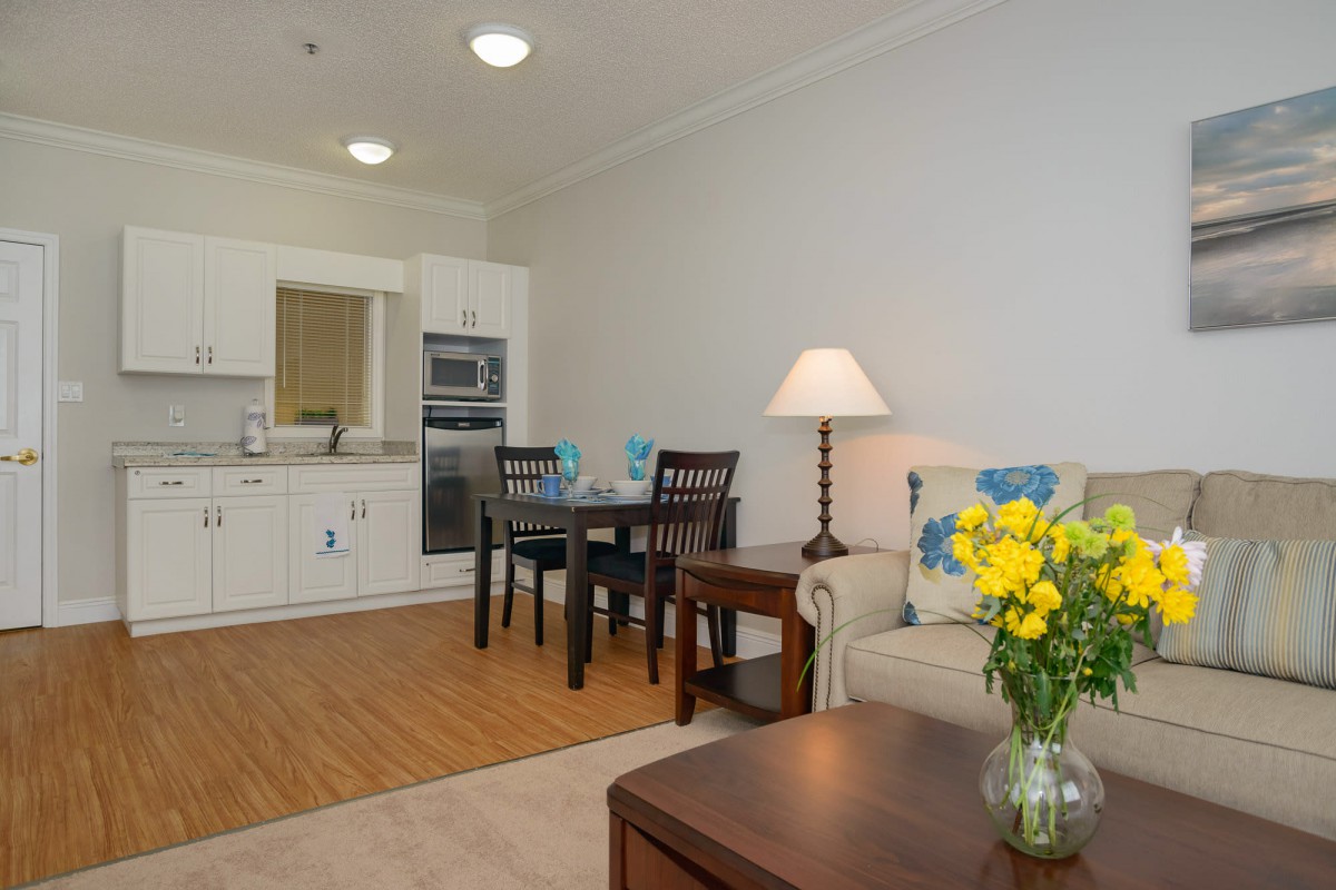 A cozy open-plan living space with a kitchen and dining area, featuring a sofa, table lamp, and a vase of yellow flowers.