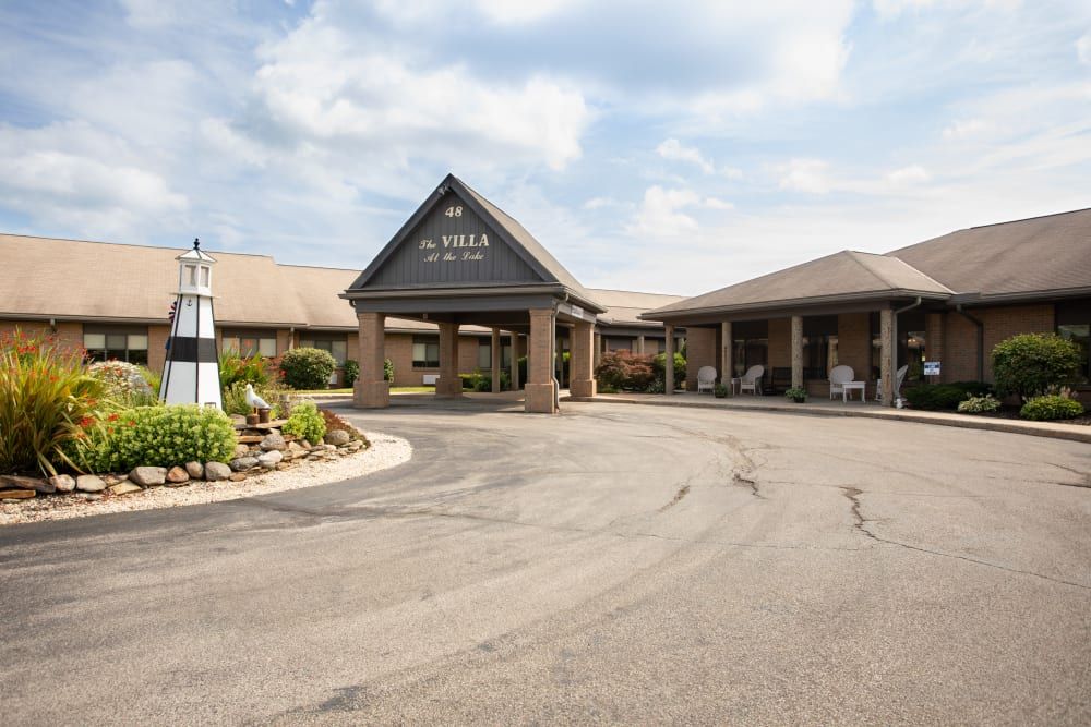 Entrance to a building with a driveway and landscaped surroundings under a cloudy sky.