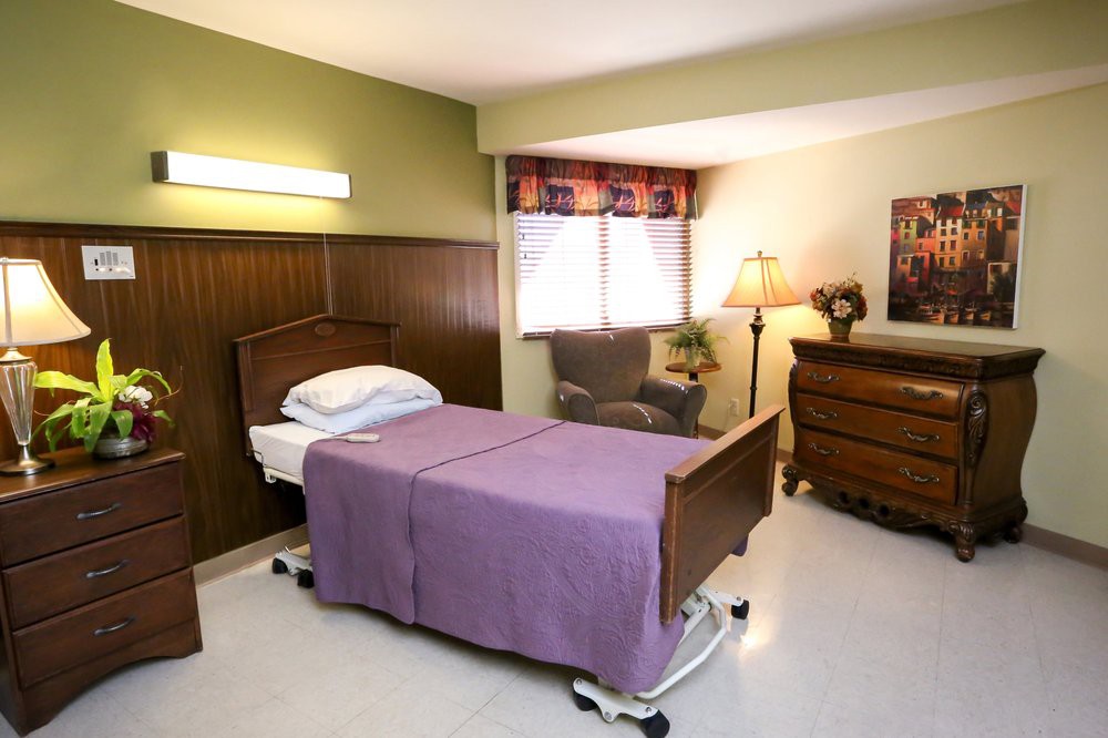 A room featuring a hospital bed with a purple blanket, next to a chair and lamp.