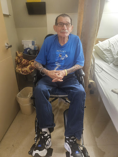 A man in a wheelchair wearing a blue shirt and displaying tattoos, seated in a cozy indoor environment.