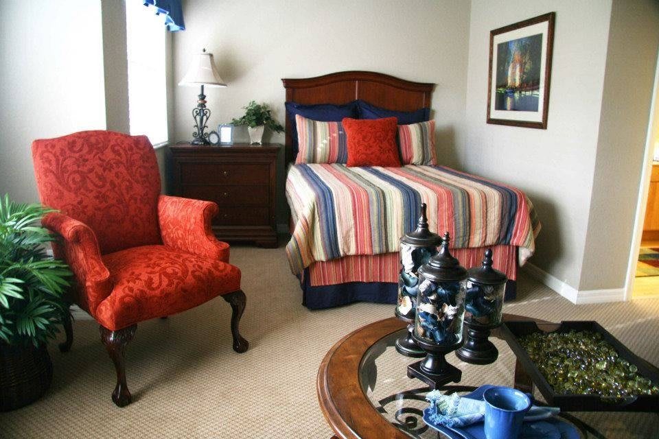 A cozy bedroom with a colorful striped bedspread and decorative pillows, an armchair, a wooden dresser, and a coffee table with decorative items.