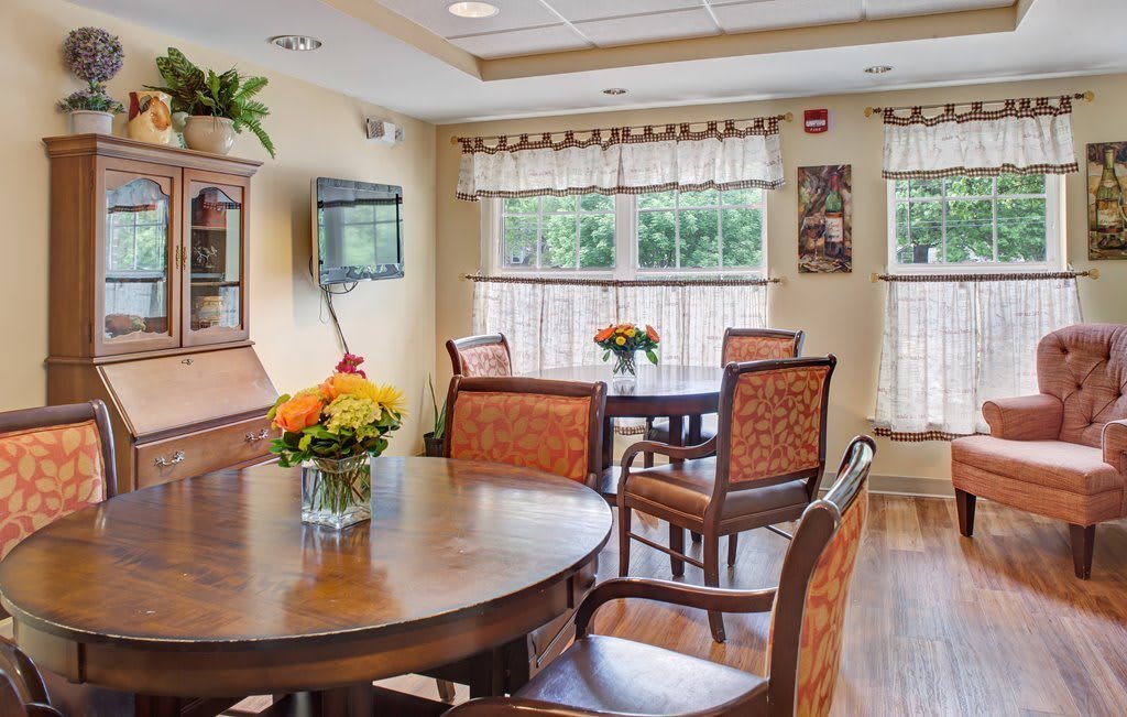 A cozy dining area with a wooden table and chairs, featuring decorative flowers and large windows.