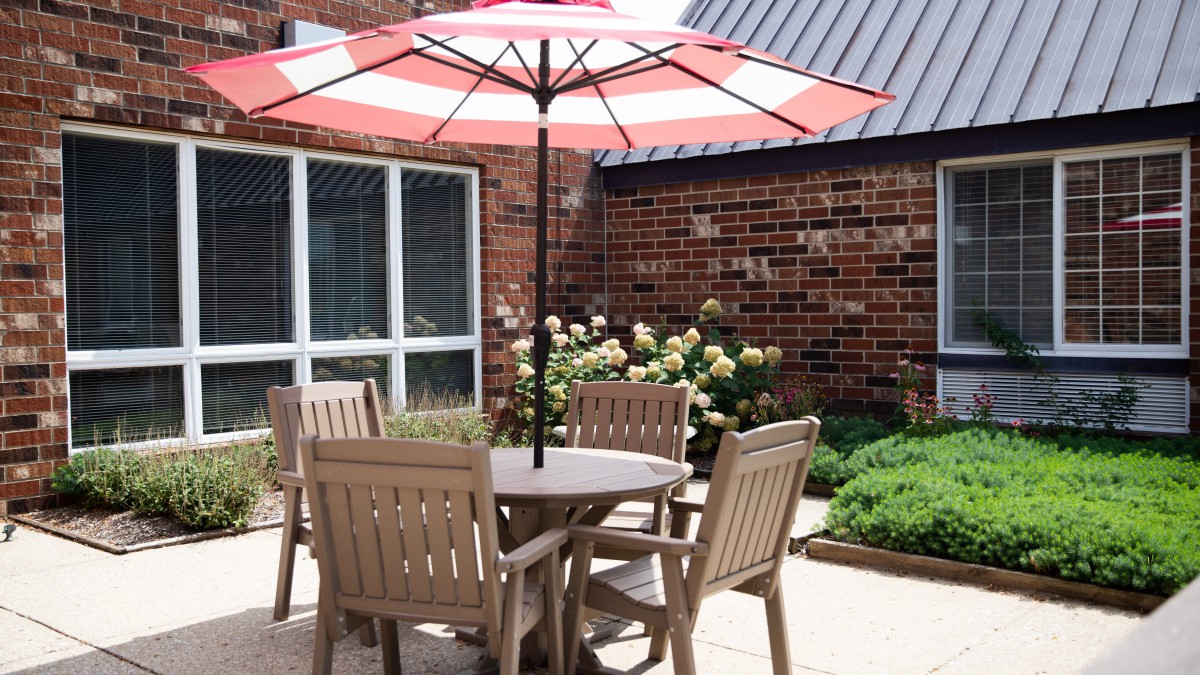 A cozy patio setup with a round table and chairs under a red and white striped umbrella, surrounded by a garden with brick walls.