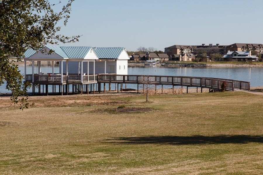 A scenic view of a lake with a wooden pier extending over the water, surrounded by grassy fields and residential buildings in the background.