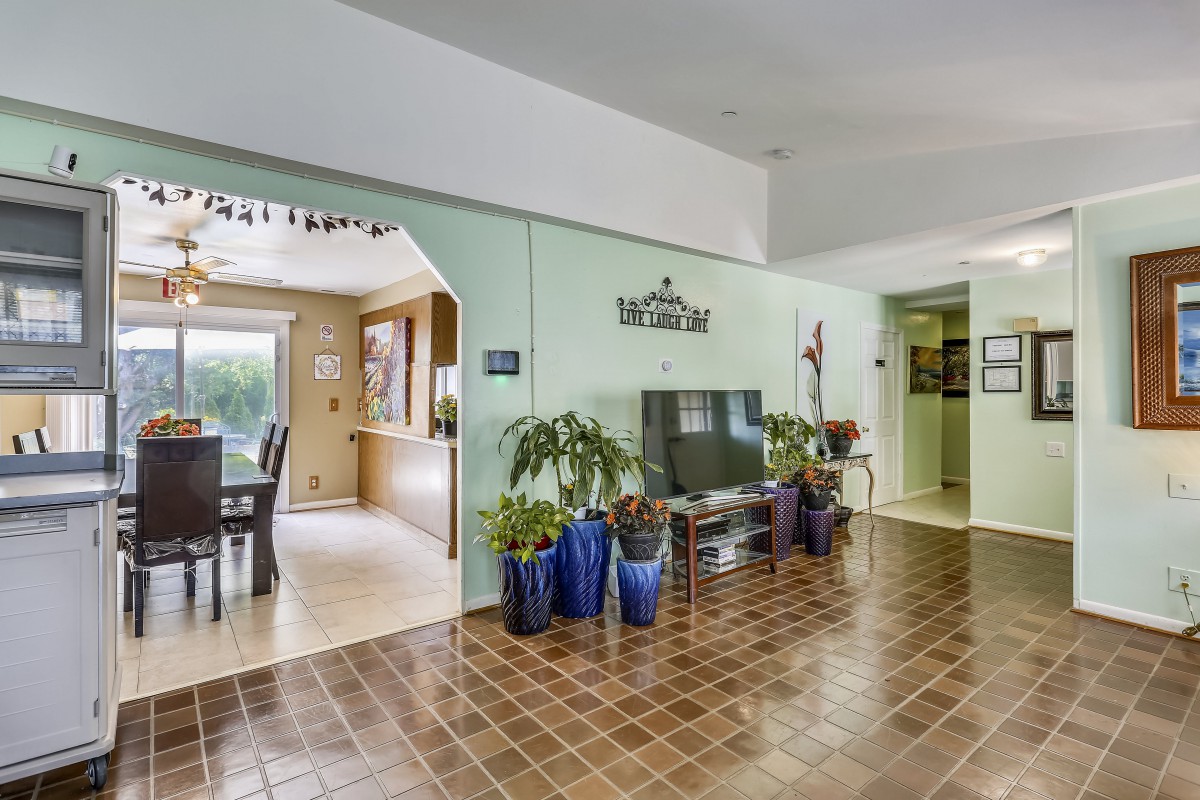 A cozy living room with tile flooring, featuring a television and potted plants, opening into a dining area.