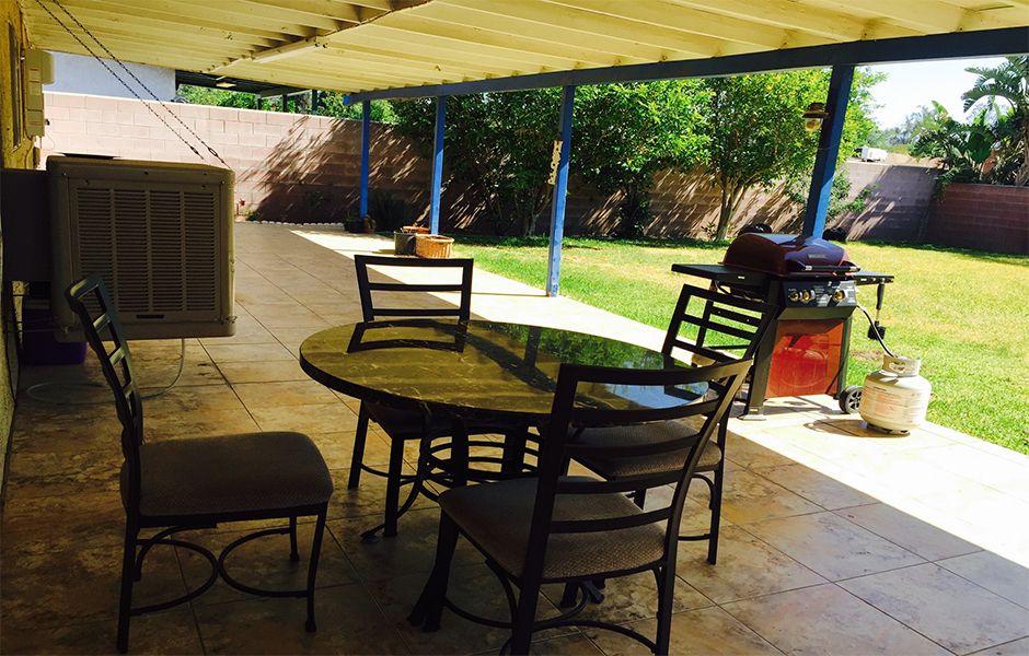 A sunny backyard patio with a round glass table and four chairs under a covered area.