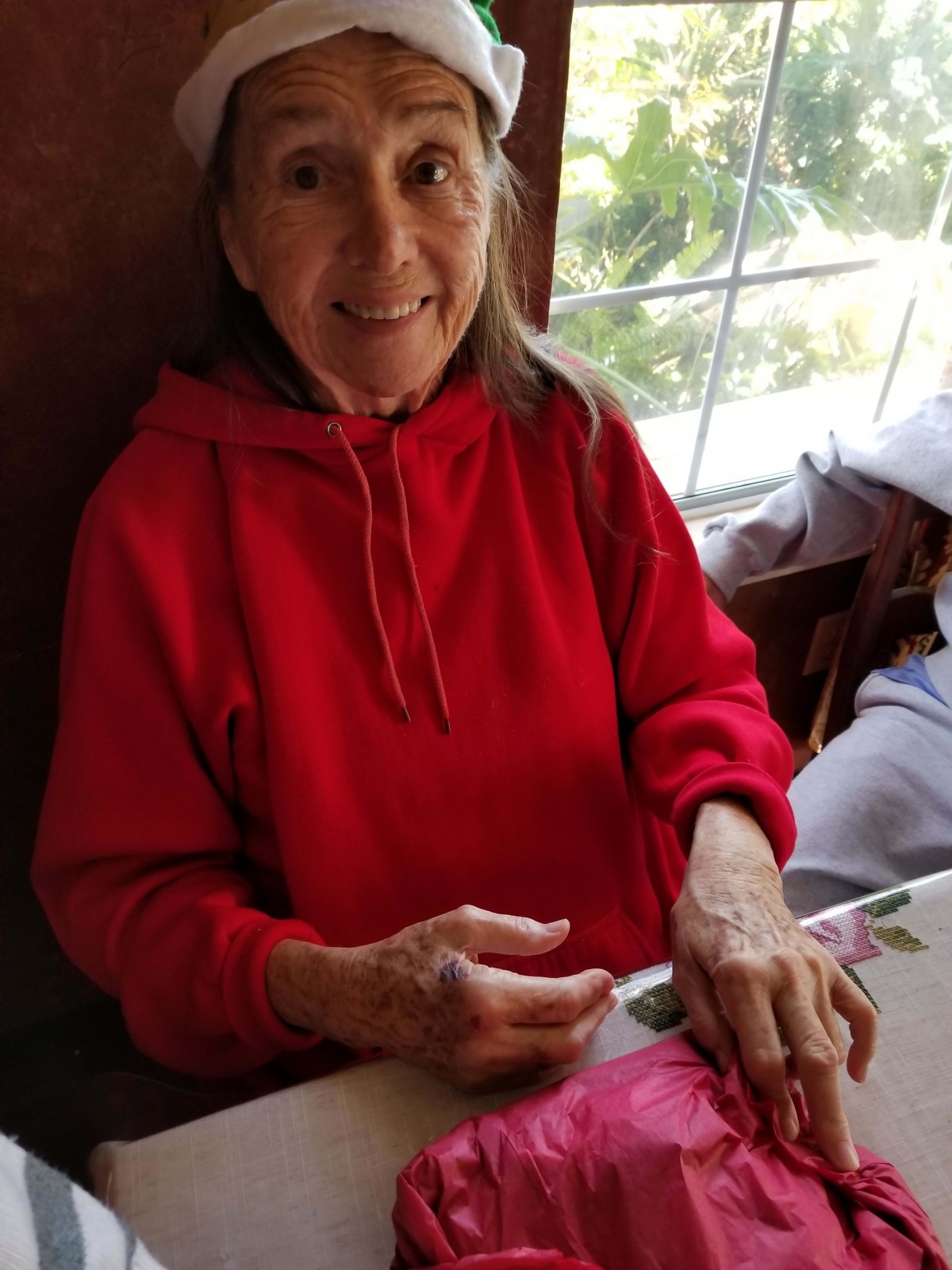 An elderly woman wearing a red hoodie is smiling while engaged in crafting indoors.