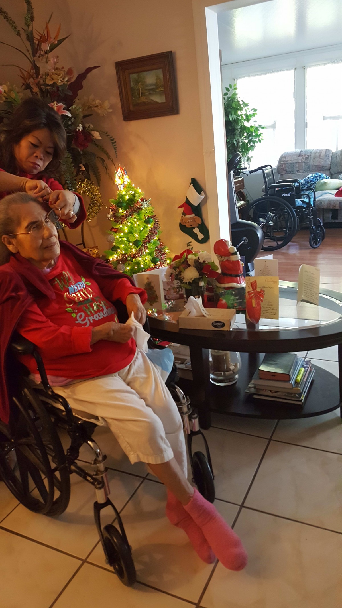 An elderly woman in a wheelchair is getting her hair done by a caregiver beside a decorated Christmas tree in a living room.