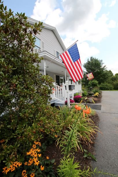 A charming house with an American flag on the porch, surrounded by lush plants and a driveway.