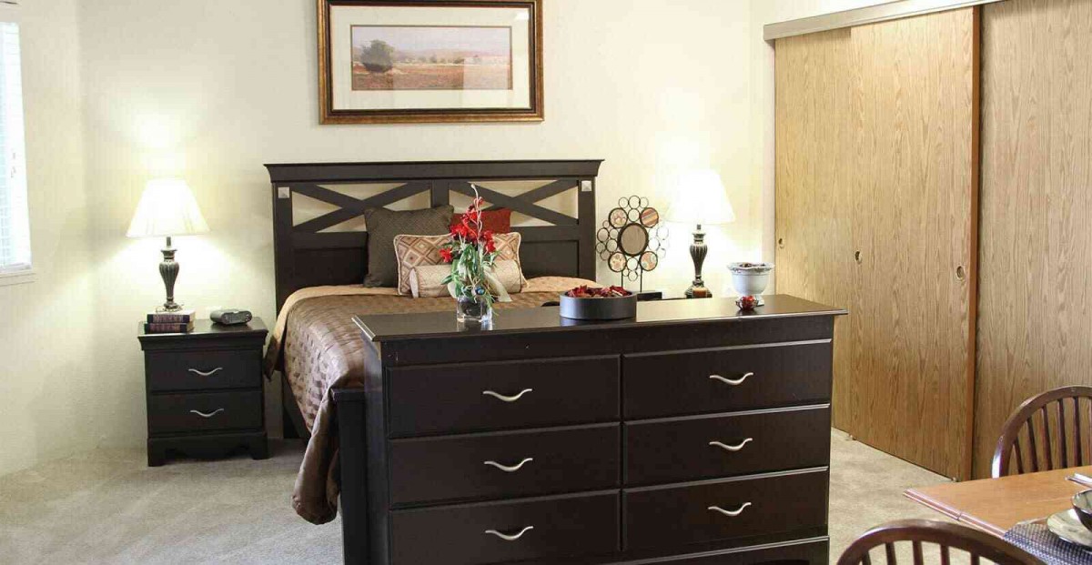 A cozy bedroom featuring a bed with a dark headboard, a large dresser, and decorative items, including a lamp and flowers.