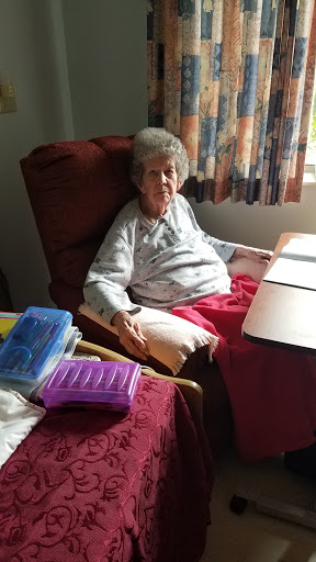 An elderly woman sitting comfortably in her armchair, surrounded by personal items in a sunlit room.