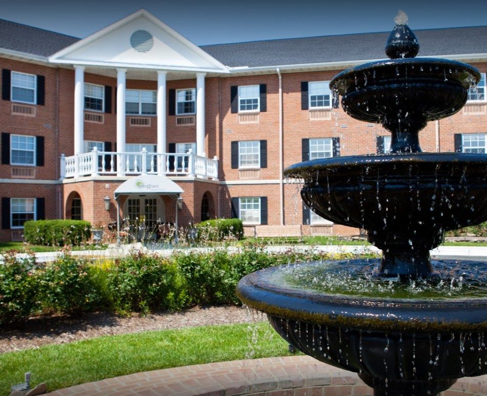 A traditional brick building with white columns stands behind a decorative fountain amidst a landscaped garden.