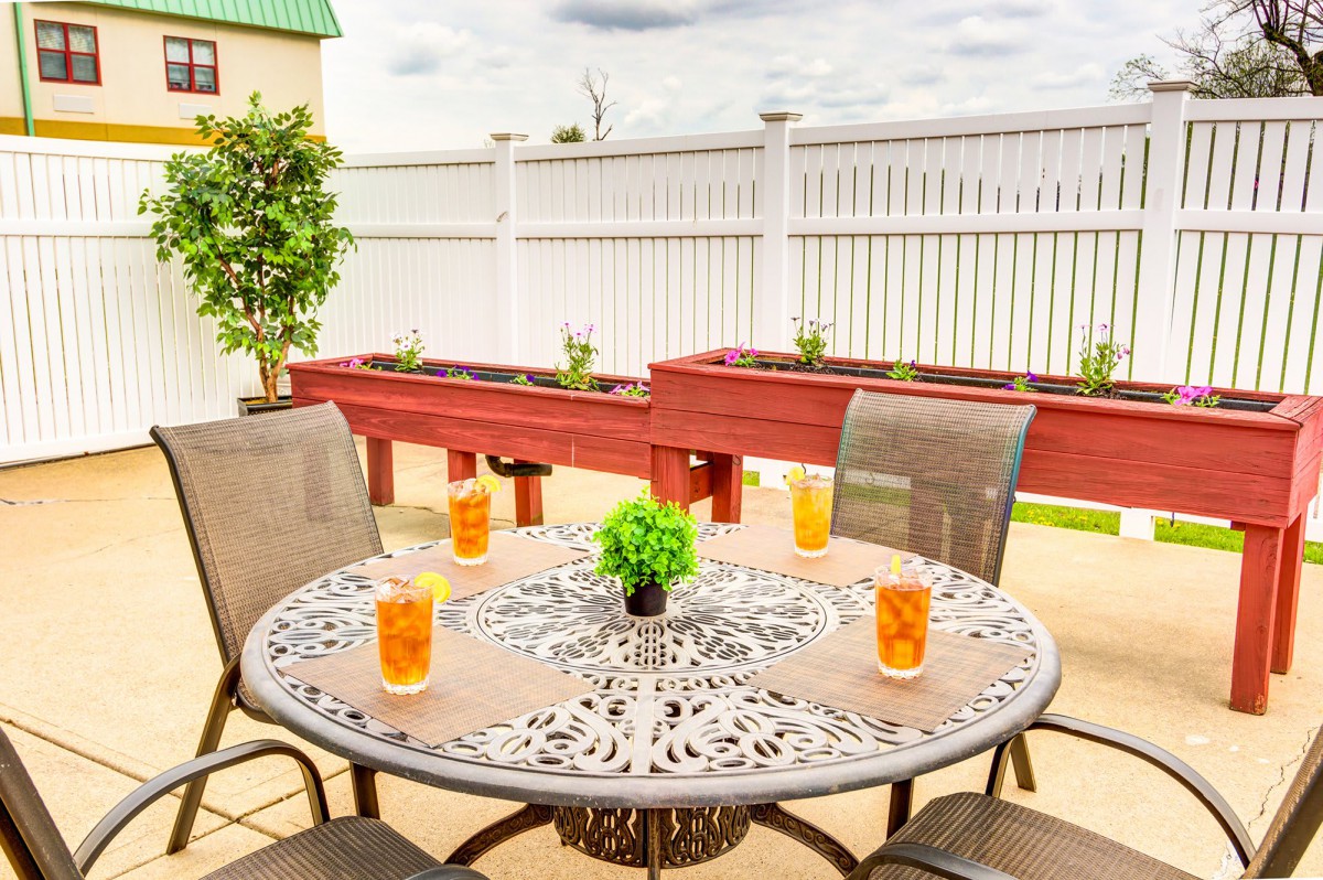 A charming patio setup with a decorative metal table, surrounded by chairs and adorned with glasses of iced tea, set against a backdrop of planter boxes and a white fence.