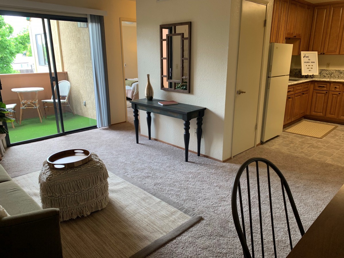 A cozy living room with carpeted flooring, featuring a table and mirror, adjacent to a kitchen and balcony.