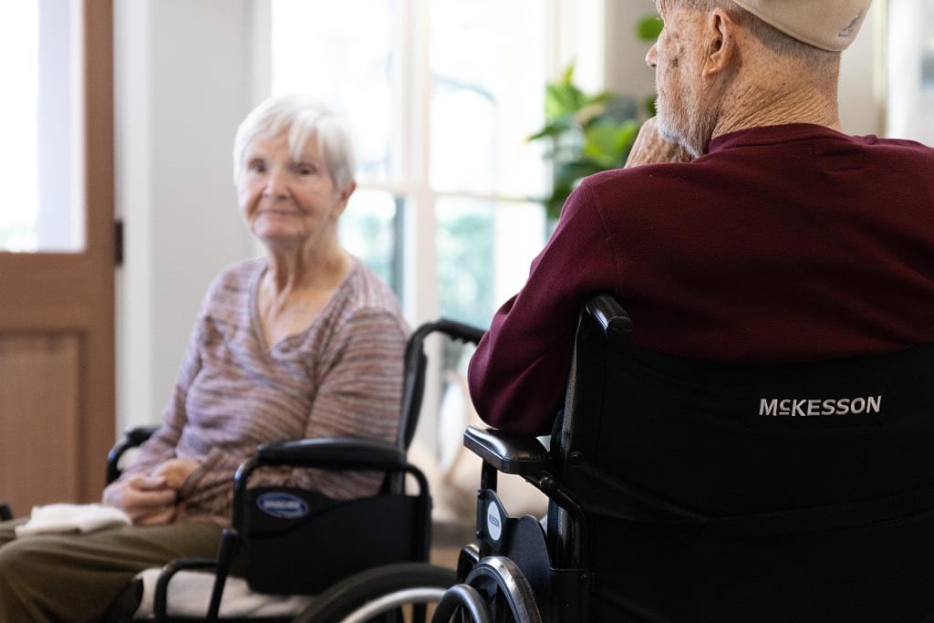 Two elderly individuals sitting in wheelchairs engage in conversation at a care facility.