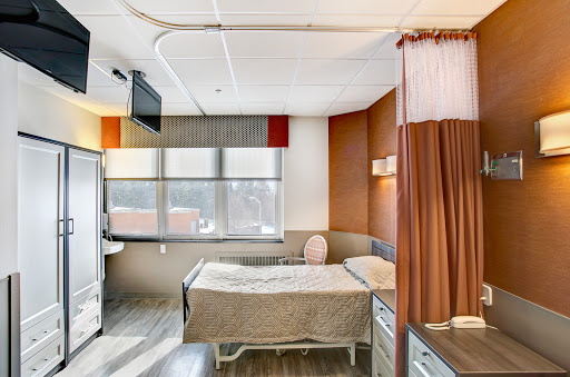 A modern hospital patient room featuring a bed, daylight from the window, and warm decor.