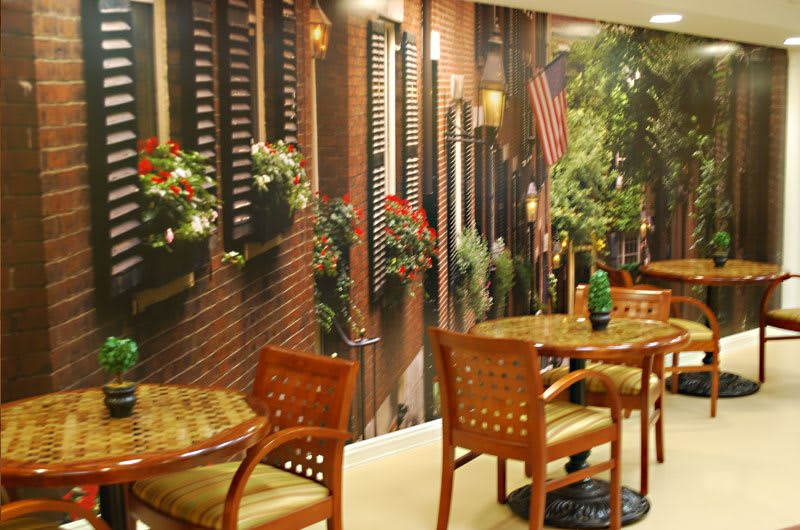 A cozy cafe interior with wooden tables and chairs, decorated with indoor plants and a mural of a street scene.
