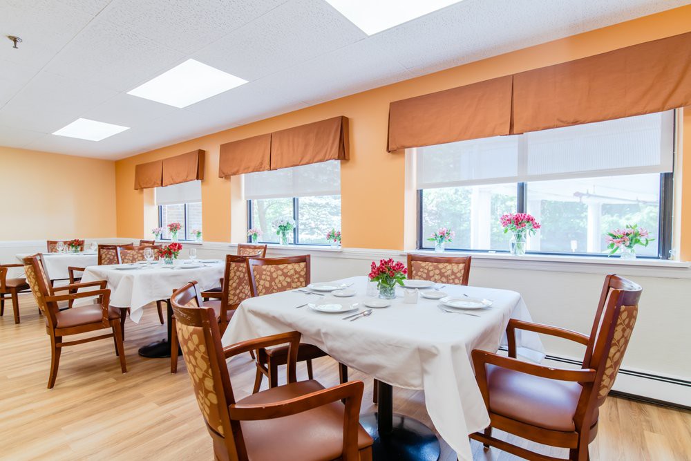 A bright dining room with tables set for a meal, featuring flowers and large windows.