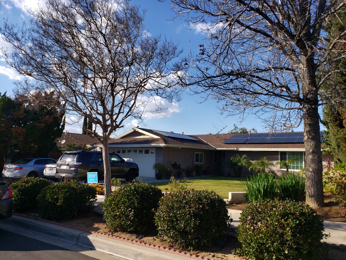 A suburban house with solar panels on the roof, cars parked in the driveway, and trees in the yard under a clear blue sky.