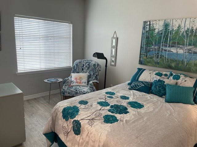 A cozy bedroom with teal and beige decor, featuring a bed, chair, window, and wall art.