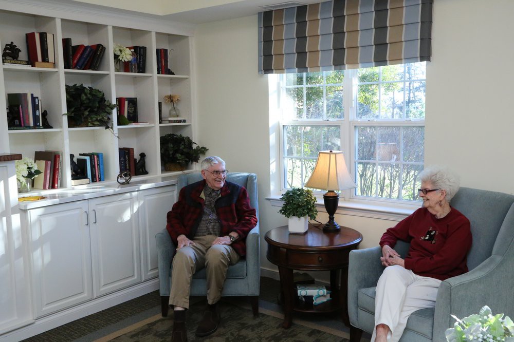 Two elderly individuals sit comfortably in a cozy living room with a bookshelf and window.
