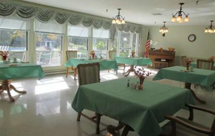 A well-lit dining room with tables covered in green tablecloths, surrounded by chairs, and large windows letting in natural light.