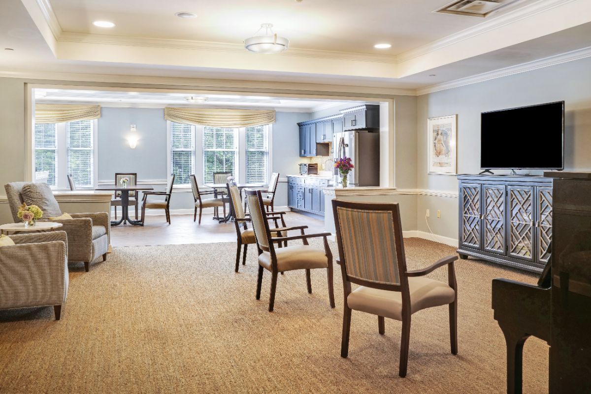 A well-lit dining and kitchen area with chairs and tables arranged on a carpeted floor.